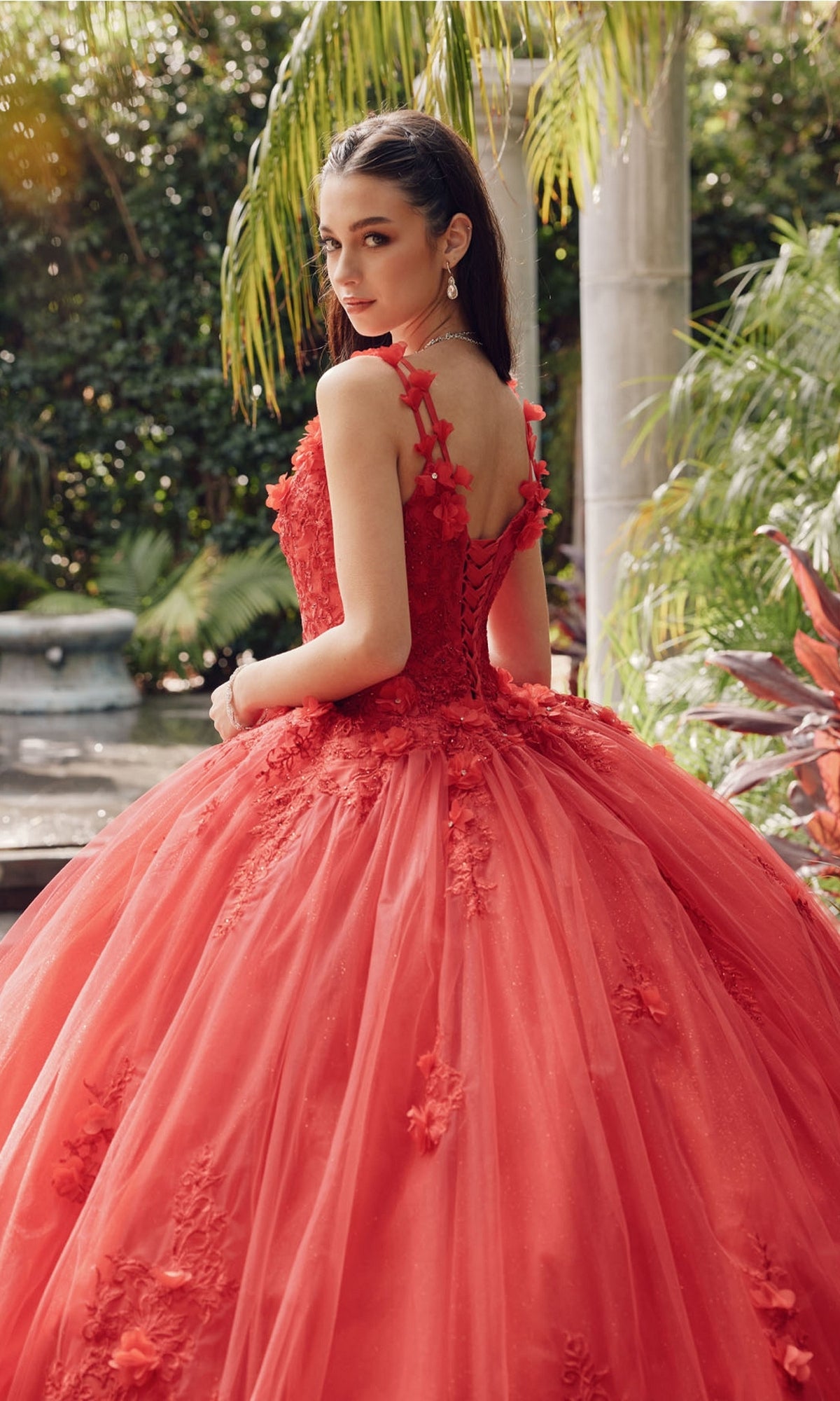 A young woman stands outdoors in the Juliet 1437R Red Ball Gown Quinceañera Dress, featuring floral details, beaded embroidery, and a lace-up back, surrounded by lush greenery and tropical plants.